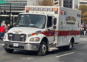 An ambulance from the Silver Spring Volunteer Fire Department drives down a city street during the day. People are visible in the background, along with buildings and trees under a clear sky.