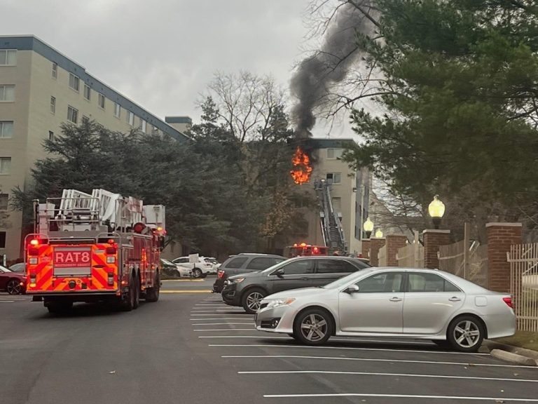 A fire truck with an extended ladder is parked in front of an apartment building with a fire and black smoke coming from a top floor. Other cars are parked along the road, and trees are visible near the building.
