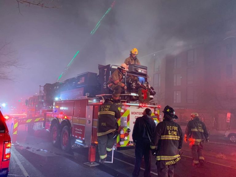 Firefighters in gear are working on a fire truck with a ladder extended towards a smoky building at night. Emergency lights illuminate the scene, and smoke fills the air.